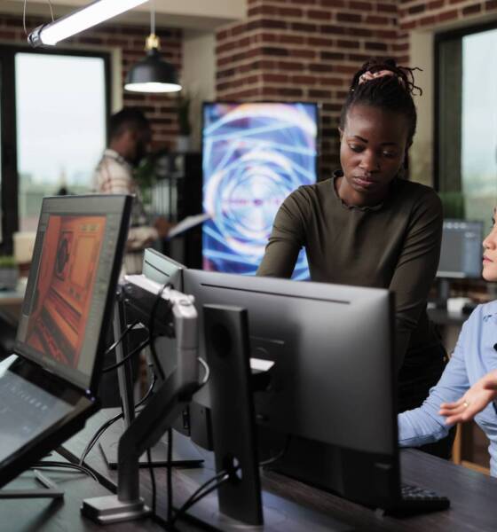 3D graphic diverse artists in office workspace conversating about design of project. Development department team leader helping coworker with software issue while working on digital art.